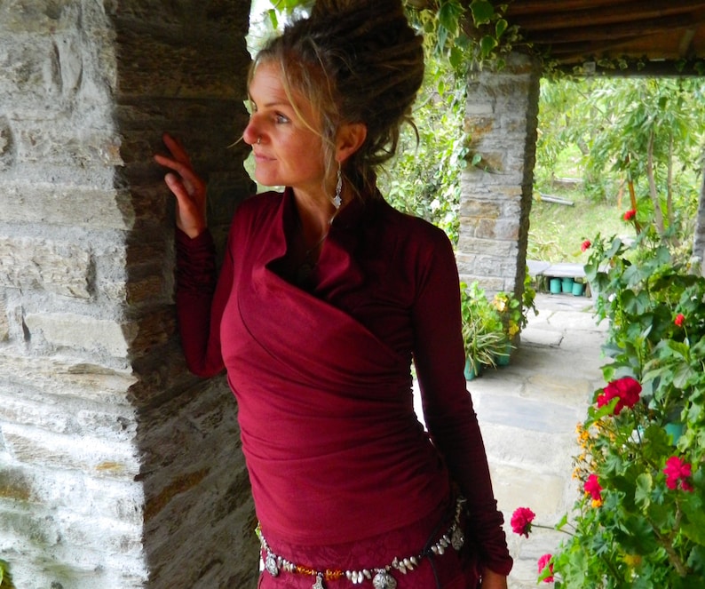 May include: A woman wearing a maroon wrap top with long sleeves and a silver belt. She is standing in front of a stone wall and looking to the right.