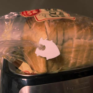 May include: A loaf of bread in a clear plastic bag, secured with a white plastic bread clip. The bread clip is shaped like a stylized fish. The bread is light brown and the bag is partially visible.