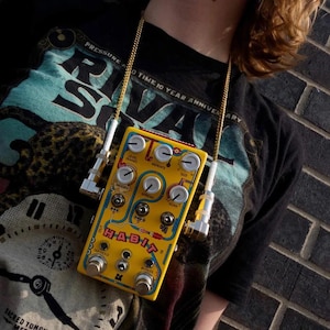 Op de afbeelding: Een gele elektronische effectpedaal met het woord "HABIT" erop, gedragen als ketting. Het pedaal heeft diverse knoppen, schakelaars en input/output jacks. Het is verbonden met een gouden ketting en witte kabels.