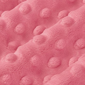 May include: Close-up of a soft, pink fabric with a textured, dotted pattern. The plush material is folded, creating shadows and highlighting the raised, rounded dots. The overall appearance suggests a cozy, tactile textile, possibly for a blanket or baby item.