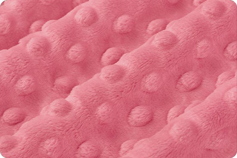May include: Close-up of a soft, pink fabric with a textured, dotted pattern. The plush material is folded, creating shadows and highlighting the raised, rounded dots. The overall appearance suggests a cozy, tactile textile, possibly for a blanket or baby item.