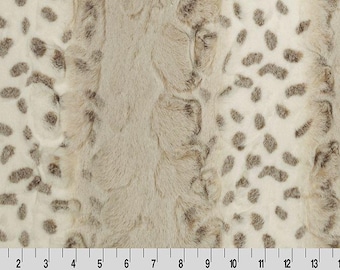 May include: A close-up of a faux fur fabric with a spotted pattern. The fabric has a cream-colored base with irregular brown spots. The fur has a soft, textured appearance, and a ruler is at the bottom.