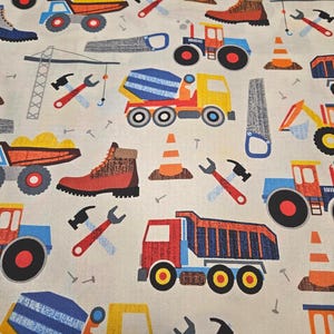 May include: A cream-colored fabric featuring a repeating pattern of construction vehicles, tools, and work boots. The design includes dump trucks, cement mixers, tractors, and orange traffic cones. Hammers, wrenches, and saws are also scattered across the fabric.