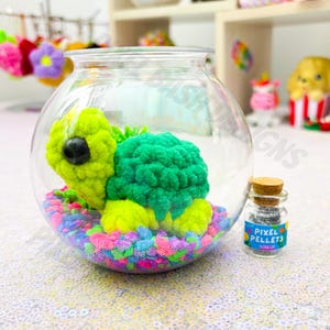 May include: A clear glass fishbowl contains a crocheted turtle with a green shell and yellow head and feet, resting on colorful gravel. A small glass vial with a cork stopper reads "PIXEL PELLETS". The background includes other decorative items.