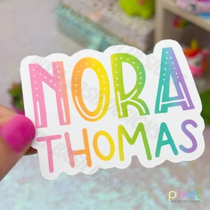 May include: A white sticker with the name "NORA THOMAS" in a rainbow gradient. The letters are outlined and filled with a gradient of pink, orange, yellow, green, and blue. The sticker has a rounded rectangular shape.