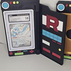 Puede incluir: Soporte para cartas impreso en 3D, negro y dorado, con una carta Pokémon calificada. La carta es una Gardevoir ex, calificada GEM MINT 10 por CGC. El soporte tiene un diseño del Team Rocket en la cubierta interior, con una gran "R" roja y detalles azules y blancos.