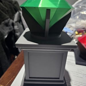 Op de afbeelding: Een 3D-geprint decoratief object. Het heeft een grote, gefacetteerde groene edelsteen op een zwarte basis, ondersteund door een zwarte structuur. De edelsteen staat op een grijze sokkel met een rechthoekig ontwerp. Het is waarschijnlijk een decoratief item.
