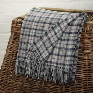 Peut inclure: Une couverture à carreaux grise, bleue et verte avec des franges. La couverture est drapée sur un panier en osier. Le motif à carreaux présente un design quadrillé avec différentes nuances de gris, de bleu et de vert. La frange est un mélange de gris et de blanc.