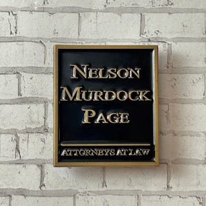 May include: A rectangular black sign with gold lettering that reads "NELSON MURDOCK PAGE ATTORNEYS AT LAW." The sign is framed in gold and mounted on a white brick wall. The sign is likely for a law firm.