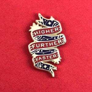 May include: Gold enamel pin featuring the words "HIGHER", "FURTHER", and "FASTER" on red banners. The banners have a navy blue background with white star and dot designs. The pin is set against a red backdrop.