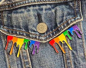 Pode incluir: Um broche de esmalte arco-íris em forma de tinta pingando em uma jaqueta de jeans. O broche está preso ao bolso da jaqueta.