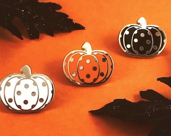 May include: Three enamel pins shaped like pumpkins with white, black, and orange polka dots. The pins are on a bright orange background.