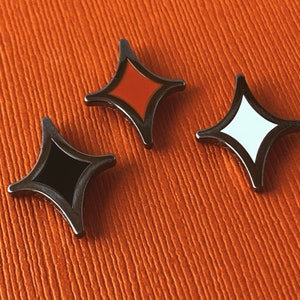 May include: Three diamond-shaped enamel pins with a black outline and a metallic silver finish. One pin is black, one is red, and one is white. The pins are displayed on an orange textured surface.