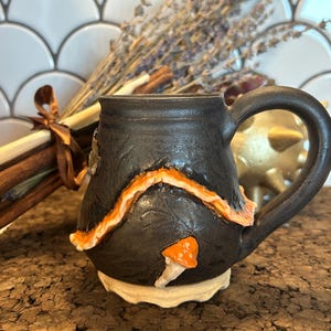 May include: A black ceramic mug with an orange and white decorative design. The mug has a large handle and a mushroom accent. Dried lavender and cinnamon sticks are in the background. The mug sits on a cork surface.