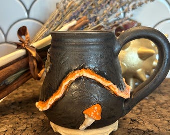 May include: A black ceramic mug with an orange and white decorative design. The mug has a large handle and a mushroom accent. Dried lavender and cinnamon sticks are in the background. The mug sits on a cork surface.