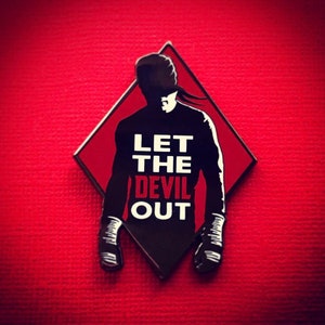 Pode incluir: Um alfinete com a silhueta preta de uma figura e o texto "LET THE DEVIL OUT" sobre um losango vermelho. O alfinete está sobre uma superfície vermelha texturizada.