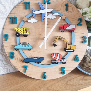 May include: A wooden clock with a blue and white airplane, a yellow dump truck, a blue car, a blue race car, a yellow excavator, and a concrete mixer truck. The clock face has numbers 1 through 12 in teal.