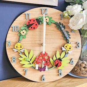 May include: A wooden clock with a whimsical garden theme. The clock face features a green wreath with ladybugs, bees, mushrooms, and a caterpillar. The numbers are in a light gray color.