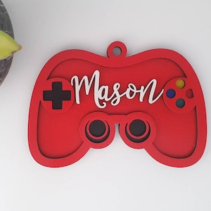 May include: A red wooden controller-shaped ornament with the name "Mason" in white script. The controller has black and multi-coloured buttons and a loop for hanging. A decorative item for gamers.
