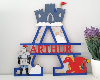 May include: A blue and white wooden letter A with a castle on top, a knight on the left side, and a red dragon on the right side. The name "ARTHUR" is written in red letters across the middle of the letter.