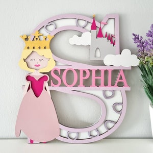 May include: A pink wooden letter S with a princess and castle design. The letter is decorated with hearts and clouds. The name "Sophia" is written in pink letters on the letter.