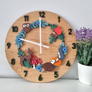 May include: A wooden clock with a brown background and black numbers. The clock face features a wreath of green leaves, blue flowers, and various animal figures, including an owl, a deer, a hedgehog, a squirrel, and a fox. The clock hands are white.