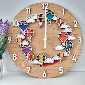 May include: A wooden clock with a light brown background and white numbers. The clock face features a design of hot air balloons in various colours, including red, blue, green, and purple. The balloons have white outlines and are decorated with white strings.