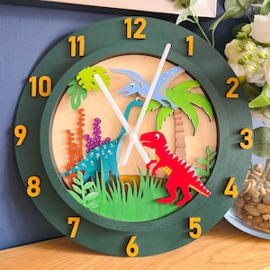 May include: A round wall clock with a dark green frame and yellow numbers from 1 to 12. The clock face features a layered wooden scene with a red T-Rex, a blue long-necked dinosaur, a flying pterodactyl, green palm trees, and plants.