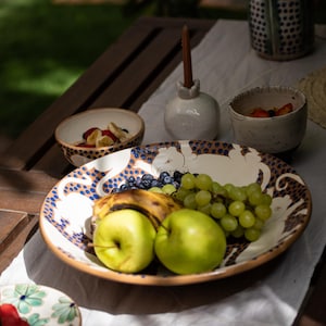 Puede incluir: Un plato de servir de cerámica blanca con un patrón de lunares azules y marrones y un diseño de gato blanco. El plato está lleno de uvas verdes, manzanas amarillas, un plátano y arándanos.