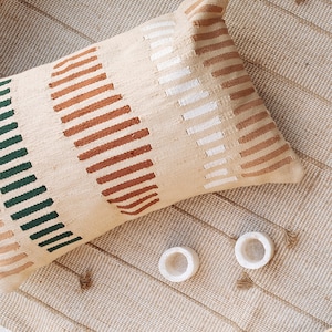 May include: A beige throw pillow with a geometric pattern in shades of brown, green, and white. The pillow is lying on a woven rug.