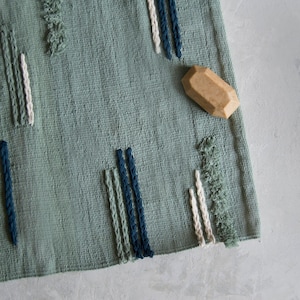 Puede incluir: Una alfombra tejida verde con detalles de flecos azules, blancos y verdes. Una pequeña forma geométrica de madera marrón claro está encima de la alfombra.