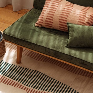Peut inclure: Un canapé en velours vert avec un cadre en bois et trois coussins. Les coussins sont de couleur rose clair avec des rayures brunes, un velours vert foncé et un velours brun. Le canapé est posé sur un tapis crème à rayures brunes et vertes.