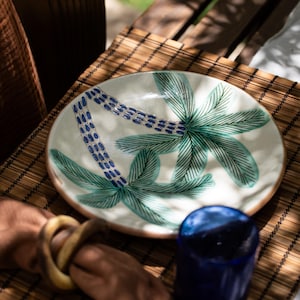 May include: A white ceramic plate with a green and blue palm tree design. The plate is sitting on a woven bamboo placemat.