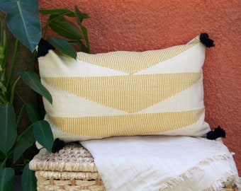 May include: Cream rectangular cushion with a yellow geometric design and black tassels. The cushion is on a white blanket, with a woven basket and a green plant in the background. The background is a terracotta wall.