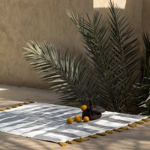 May include: A white and black striped rug with yellow tassels. The rug is laid out on a concrete patio with a palm tree in the background. A small wicker basket with oranges is on the rug.