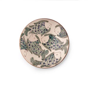 May include: A round ceramic plate with a cream-colored base, featuring a hand-painted design of six stylized fish. The fish are in shades of blue and green, with a brown rim. The plate is a decorative item.