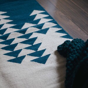 Op de afbeelding: Een wit tapijt met een blauw geometrisch patroon van driehoeken. Het tapijt ligt op een houten vloer en een blauw gehaakt item bevindt zich in de rechterbenedenhoek.