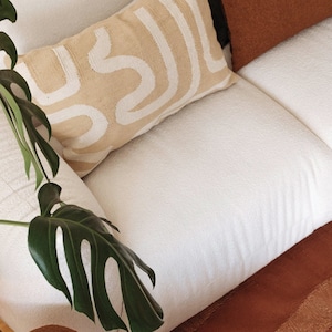 May include: A white couch with a beige pillow with a white abstract design. A green plant is in the foreground.