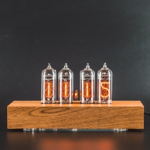 May include: A wooden clock with four clear glass tubes displaying the time as 10:15. The clock has a warm wood finish and is sitting on a black surface.