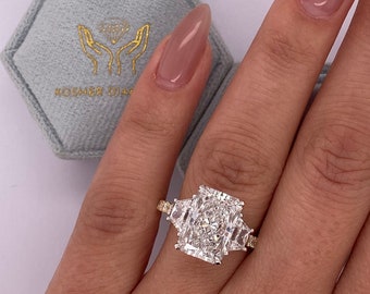 May include: A gold ring featuring a large, rectangular, clear diamond in the centre. The diamond is surrounded by smaller diamonds. The ring is worn on a person's finger.