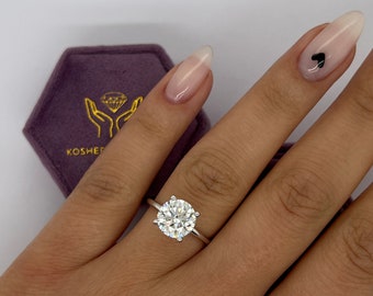 May include: A close-up of a hand wearing a silver ring with a large, round, clear diamond. The ring is on the middle finger of the hand. The hand is resting on a purple velvet box with the word "KOSHER" printed on it.