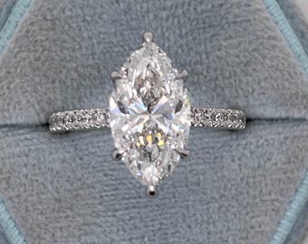Könnte beinhalten: Ein Verlobungsring mit einem Diamanten im Marquiseschliff in einem silbernen Band. Das Band ist mit kleinen Diamanten besetzt. Der Ring wird in einer hellblauen Samtringbox präsentiert.