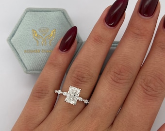 May include: A radiant rectangular diamond engagement ring with a pave setting. The ring is set in white gold and features a row of smaller diamonds on each side of the center stone.