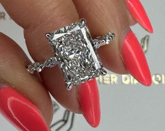 May include: A radiant-cut diamond ring with a large, rectangular center stone. The ring features a delicate band with smaller accent diamonds. The setting is a classic design, showcasing the brilliance of the diamonds. The ring is held up by a hand with bright red nail polish.