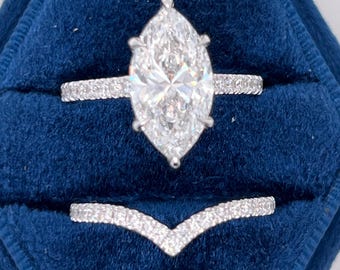 Könnte beinhalten: Ein Verlobungsring mit Diamanten im Marquise-Schliff und einem Pavé-Band, kombiniert mit einem V-förmigen Ehering, in einer tiefblauen Samt-Ringbox. Der Diamant funkelt hell und zeigt seine Facetten.