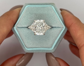 Puede incluir: Un anillo de compromiso de tres piedras con una piedra central rectangular grande y dos piedras laterales triangulares más pequeñas. El anillo está engastado en una banda de metal blanco y se muestra en una caja de anillo de terciopelo azul claro.