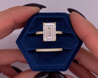 Könnte beinhalten: Ein goldener Verlobungsring mit einem großen Smaragdschliff-Diamanten und einem passenden Diamantband. Der Ring wird in einer blauen Samt-Ringbox präsentiert.