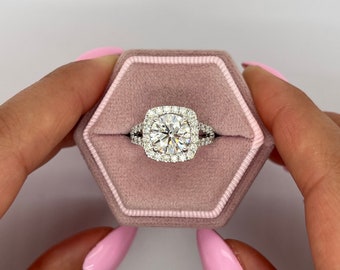 Puede incluir: Un anillo de compromiso de diamante redondo con engaste halo y una banda doble. El anillo está en una caja de anillo de terciopelo rosa.