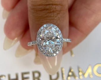 Puede incluir: Un brillante anillo de diamantes con una piedra central de talla ovalada, rodeada por un halo de diamantes más pequeños. El anillo está engastado en una banda plateada, mostrando un diseño clásico y elegante. El anillo es sostenido por una mano.