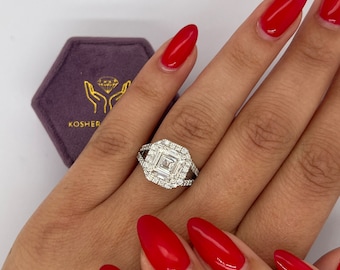 May include: A square-shaped diamond engagement ring with a halo setting. The ring is set in white gold and features a double halo of smaller diamonds. The ring is on a hand with red nail polish.